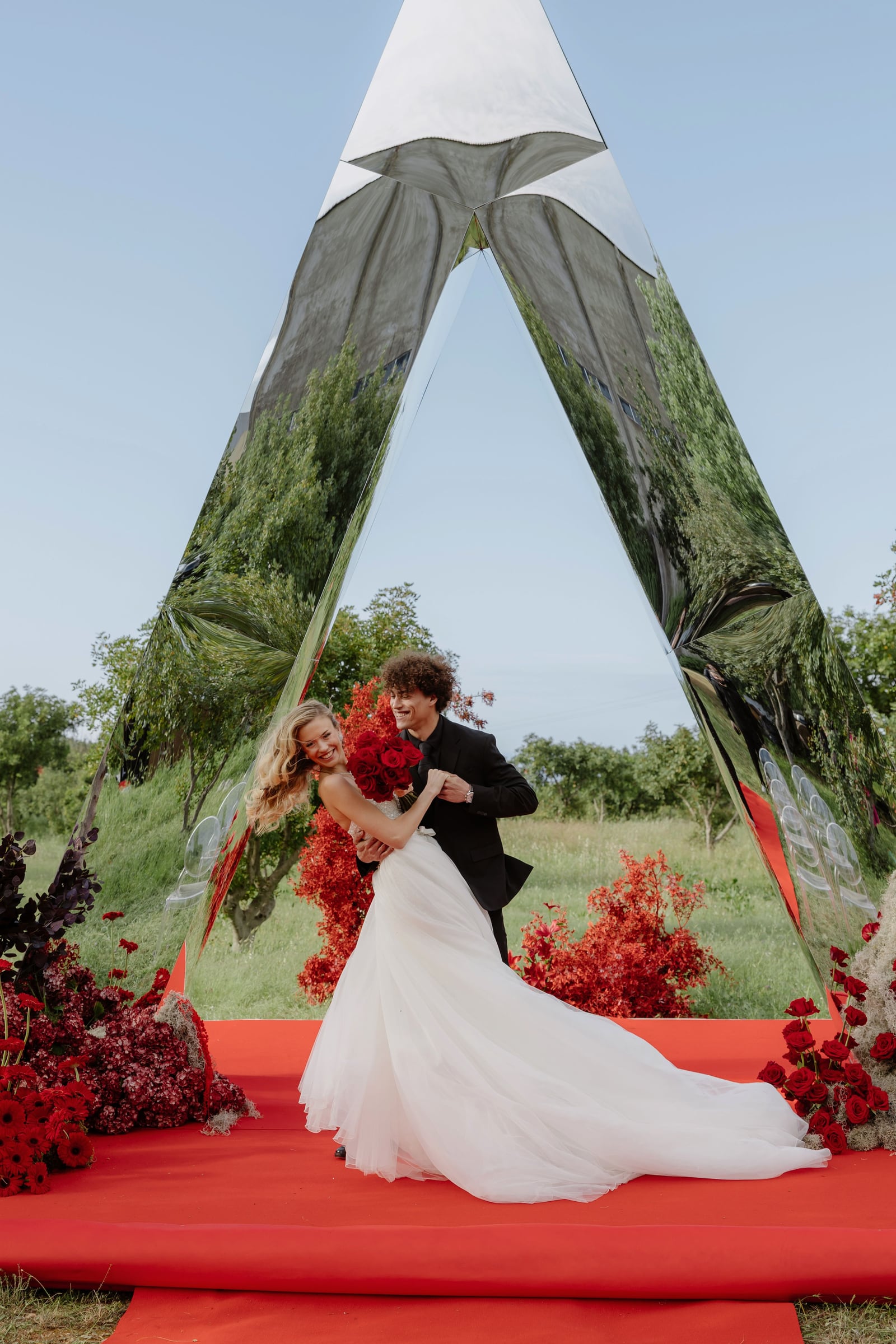 A bride in a flowing white gown laughs as a dark-suited groom lifts her slightly, both holding a bouquet of deep red roses, inside a tall triangular mirrored arch on a red carpet. Around them are red gerbera daisies, dark burgundy hydrangeas, red roses and red-leaved shrubs, with orchard trees reflected in the mirrors. This wedding flower design is for a wedding at Rethimno in Greece.