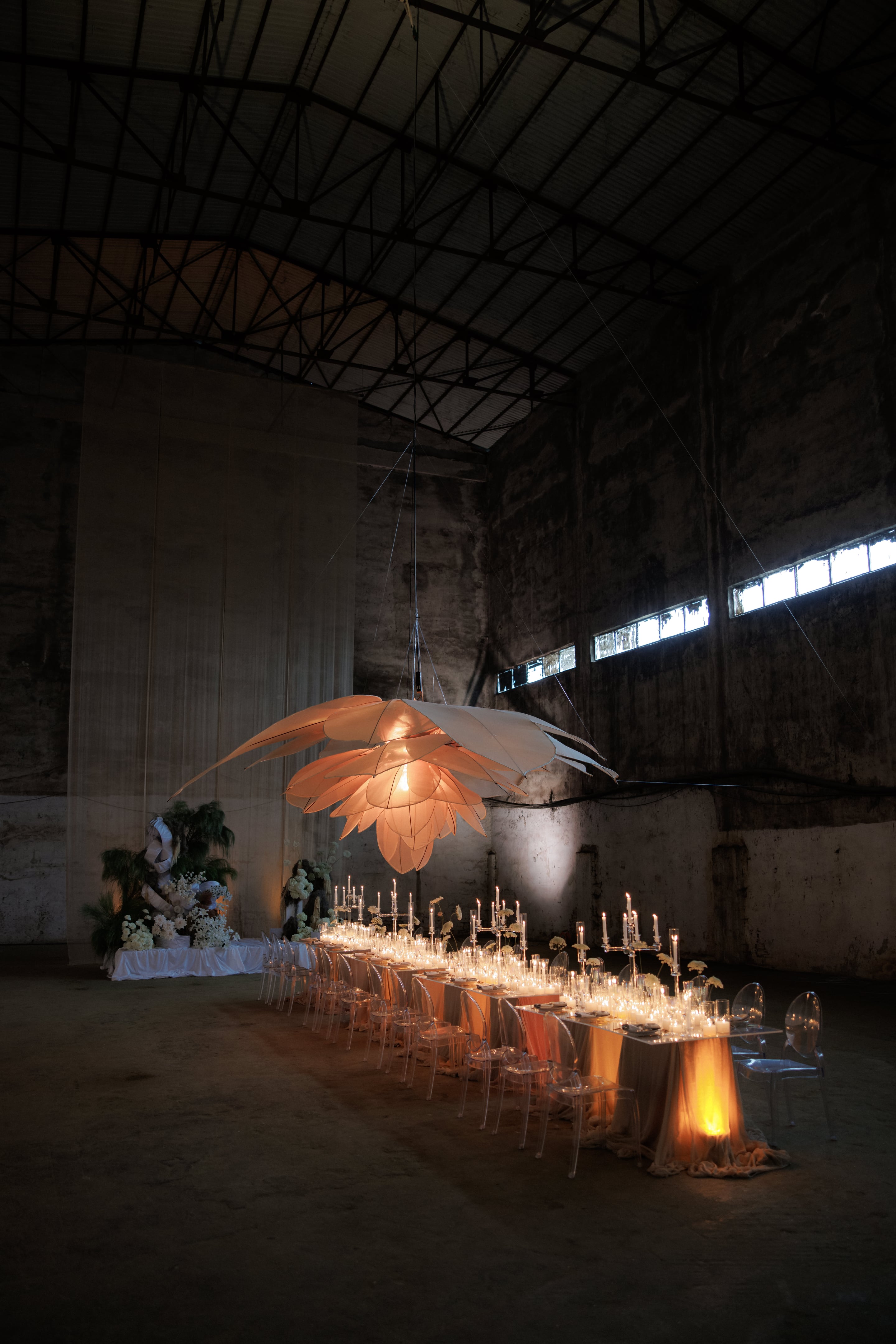 A long candlelit banquet table runs through a vast, raw concrete hall in Rethimno, Crete, Greece, set for a wedding reception. Above it hangs an enormous glowing pendant shaped like a pale peach lotus, its layered petals lit from within. Clear acrylic chairs line both sides, the table is draped in soft peach and sand-coloured fabric, and tall silver candelabras flicker among rows of candles and white blooms. At the far end, a white-draped table is dressed with cream flowers and fern-like foliage.
