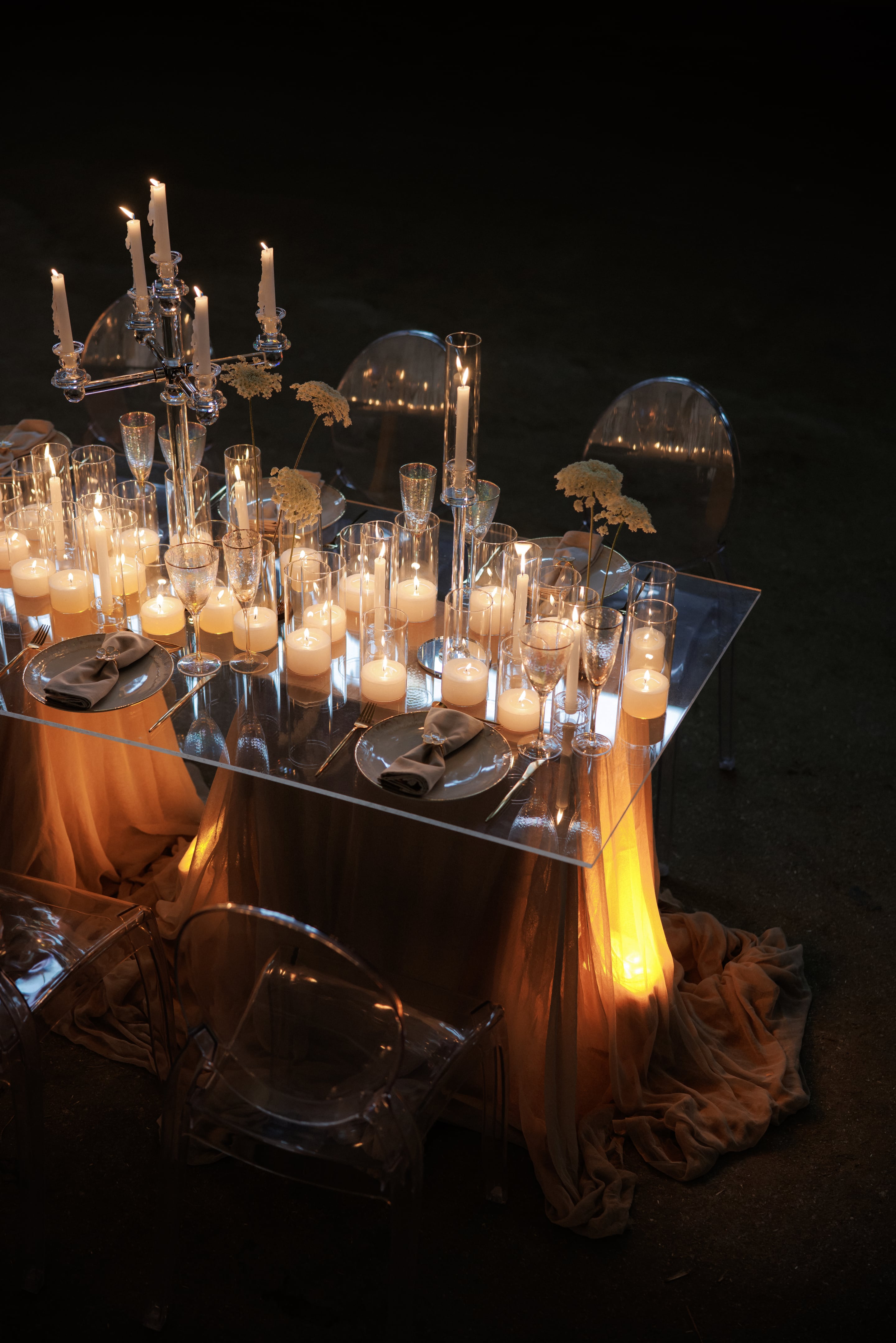 A wedding dinner table at night in Rethimno, Crete, Greece, set with a clear acrylic top and a flowing peach-toned cloth beneath it. Dozens of glowing pillar and tealight candles sit in clear glass cylinders, flanked by slender stemless glasses, grey charger plates with folded grey napkins, and delicate white umbellifer blooms rising from tall glass vases. Tall candles burn in a silver candelabra behind, and clear ghost chairs sit in the dark, giving the scene a soft, intimate, romantic glow.