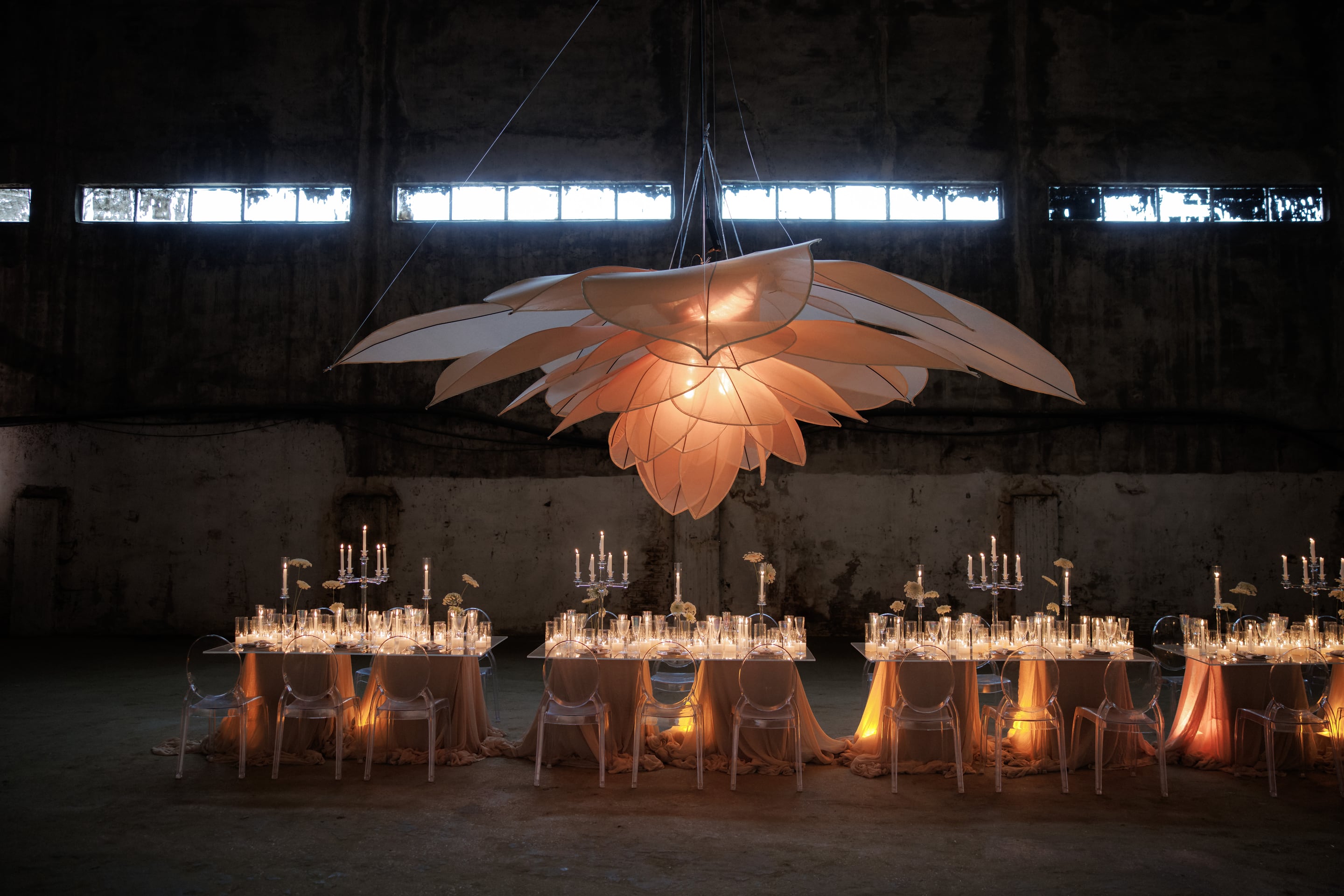 A vast, glowing petal installation hangs from the rafters like an enormous peach-toned flower, its translucent layers lit from within above three long banquet tables in a dark, weathered industrial hall. The tables are dressed with flowing blush fabric, rows of glass candleholders, tall silver candelabras and a few pale yellow blooms, with clear chairs lined up beside them. This wedding table styling is set in Rethimno, Crete, Greece.