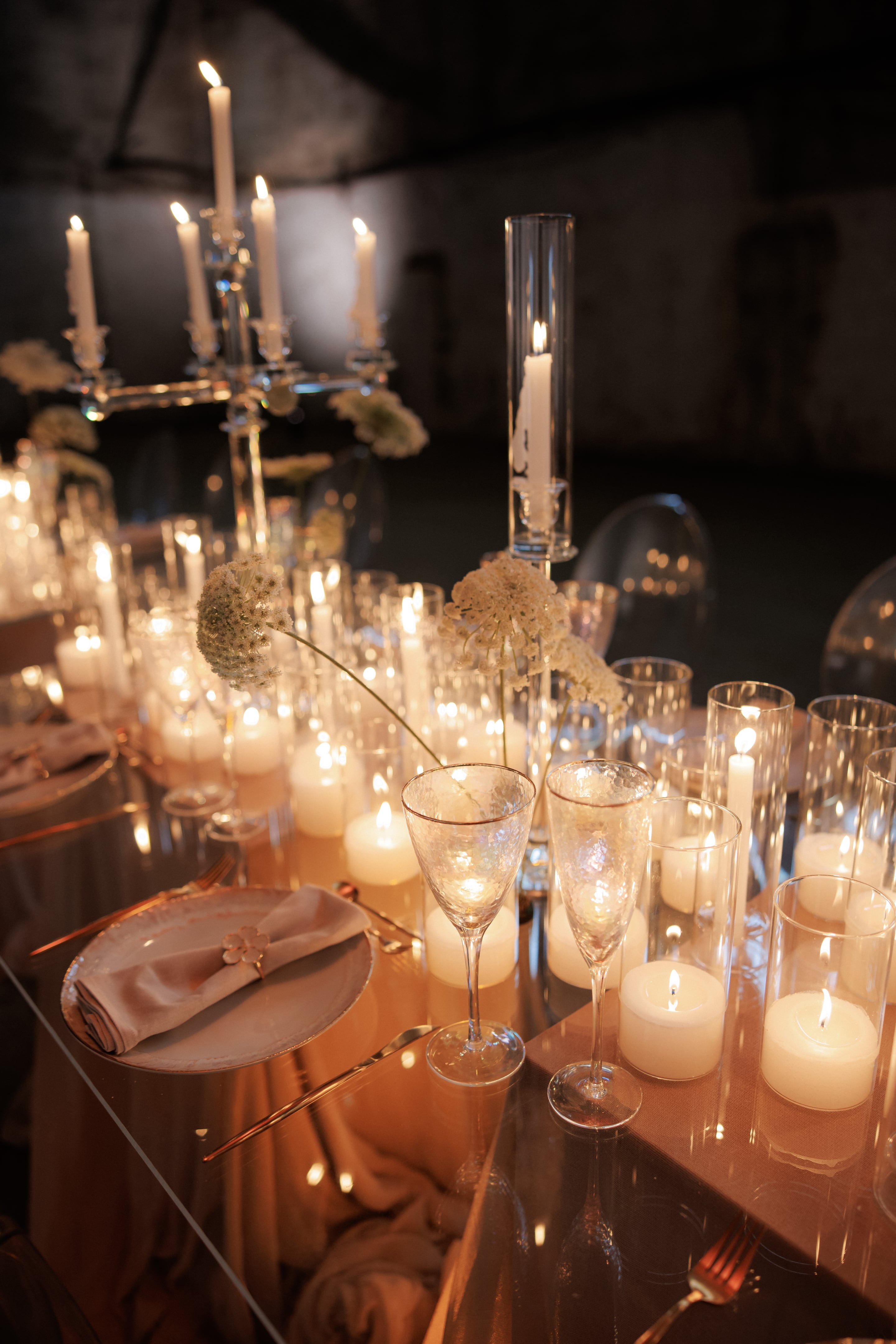 Candlelit wedding table in Rethimno, Crete, Greece, dressed with slender stems of cream-white flowers that look like Queen Anne's lace, their lacy umbels nodding over crystal flutes. Clusters of pillar candles glow inside tall glass hurricane holders, and a tall glass candle column rises at the centre, with a crystal candelabra blurred in the background. The palette is soft and warm, with blush linens and gold cutlery, and the dark room makes the flickering light feel intimate.