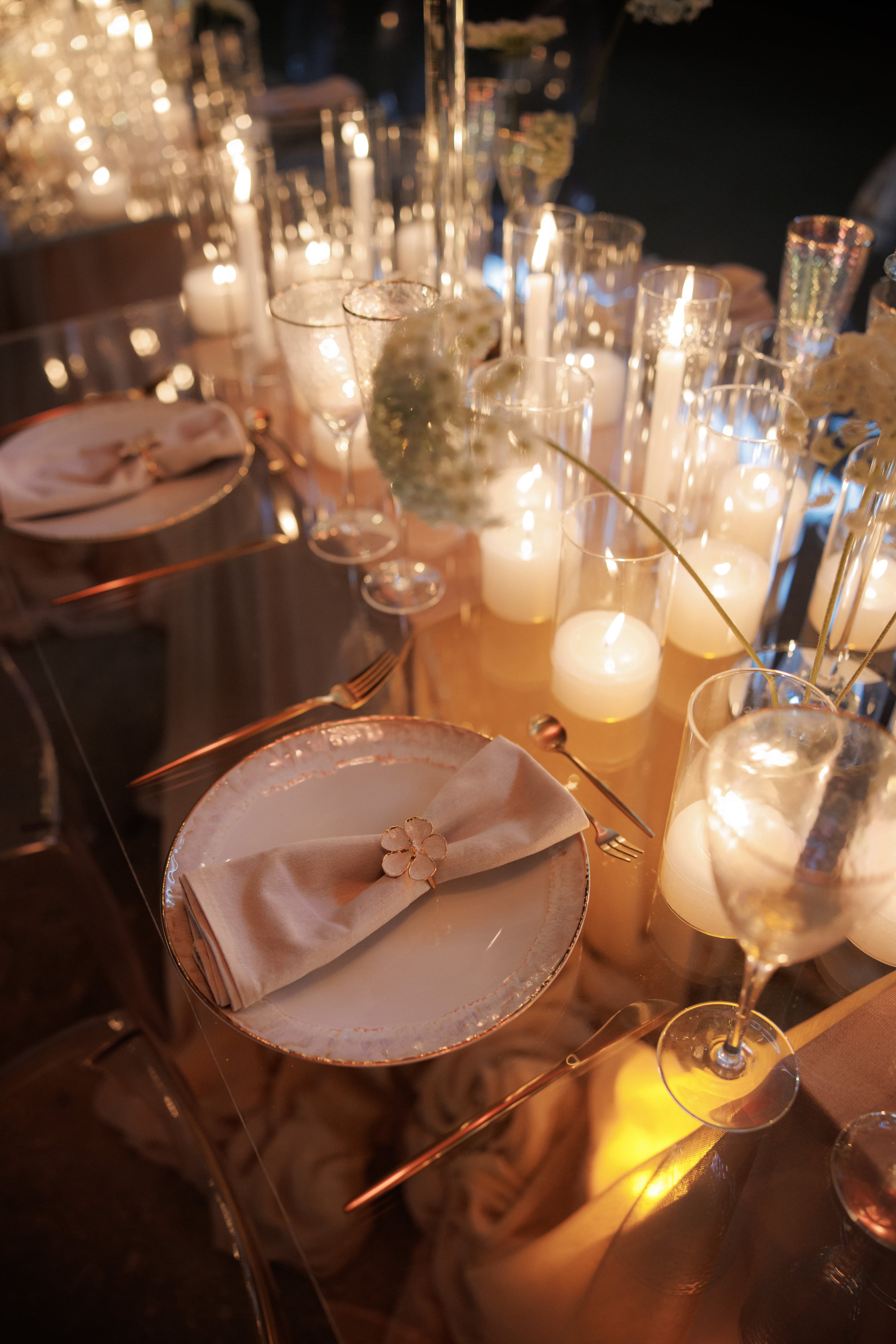 A long wedding table set in candlelight at Rethimno, Crete, Greece, with tall and pillar candles glowing inside clear glass cylinders among champagne flutes and blush-rimmed plates. Sprigs of dried white flowers, possibly gypsophila or dried Queen Anne's lace, trail through the centre, and each place setting has a pale pink napkin tied with a small gold blossom ring. The soft warm light, gold cutlery and reflective table create an intimate, elegant mood.