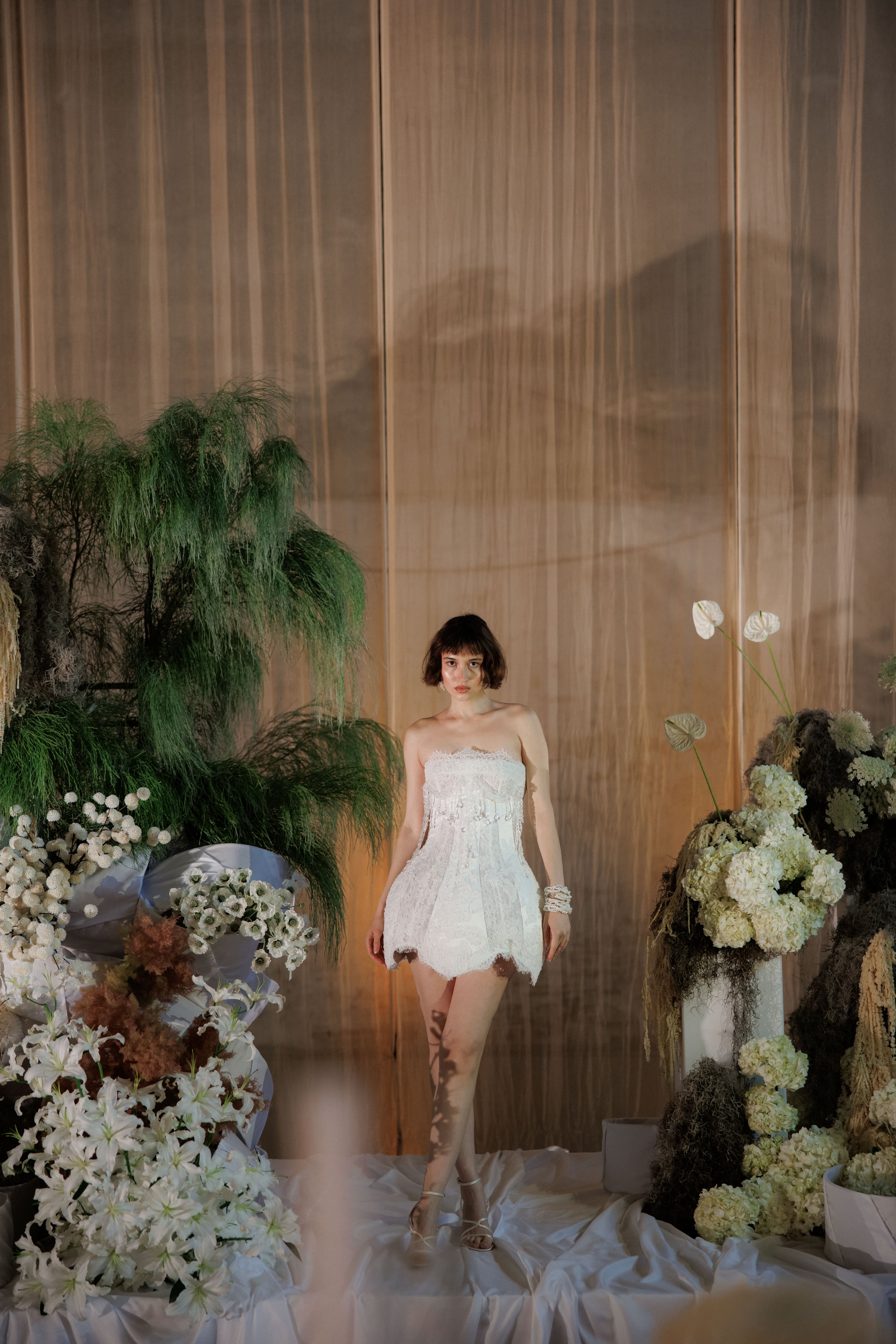 A woman with a short dark bob stands centre frame in a strapless white lace mini dress with scalloped hems and beaded fringe at the waist, set among cream and white flowers at a Rethimno, Crete, Greece wedding flower installation. Behind her and around her are white lilies, cream hydrangeas, white anthurium, and feathery green fern and foliage, with a soft gauze backdrop and a white silk-draped floor. The warm, low light gives the scene a quiet, romantic mood.