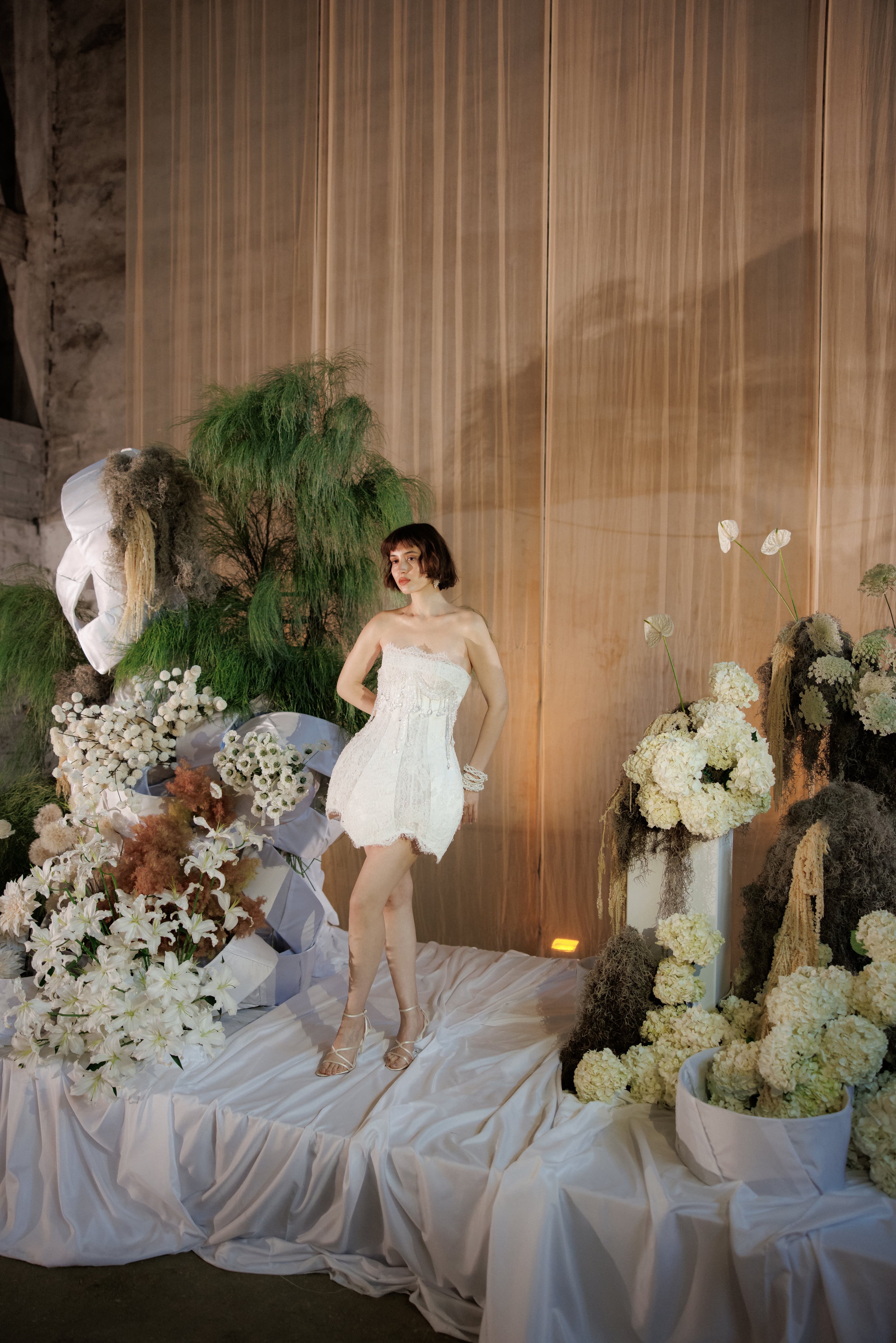 A woman with a short dark bob stands on a draped white silk cloth in a styled set of wedding flowers, wearing a strapless white lace mini dress with beaded fringe and a pearl bracelet. Around her are white lilies, white hydrangeas, anthuriums, and feathery grasses in grey and green, with a large sculptural white bloom at the left, all set against warm wooden panels. This is an editorial wedding floral display from The Extravaganza Flower, shot at a venue in Rethimno, Crete, Greece.