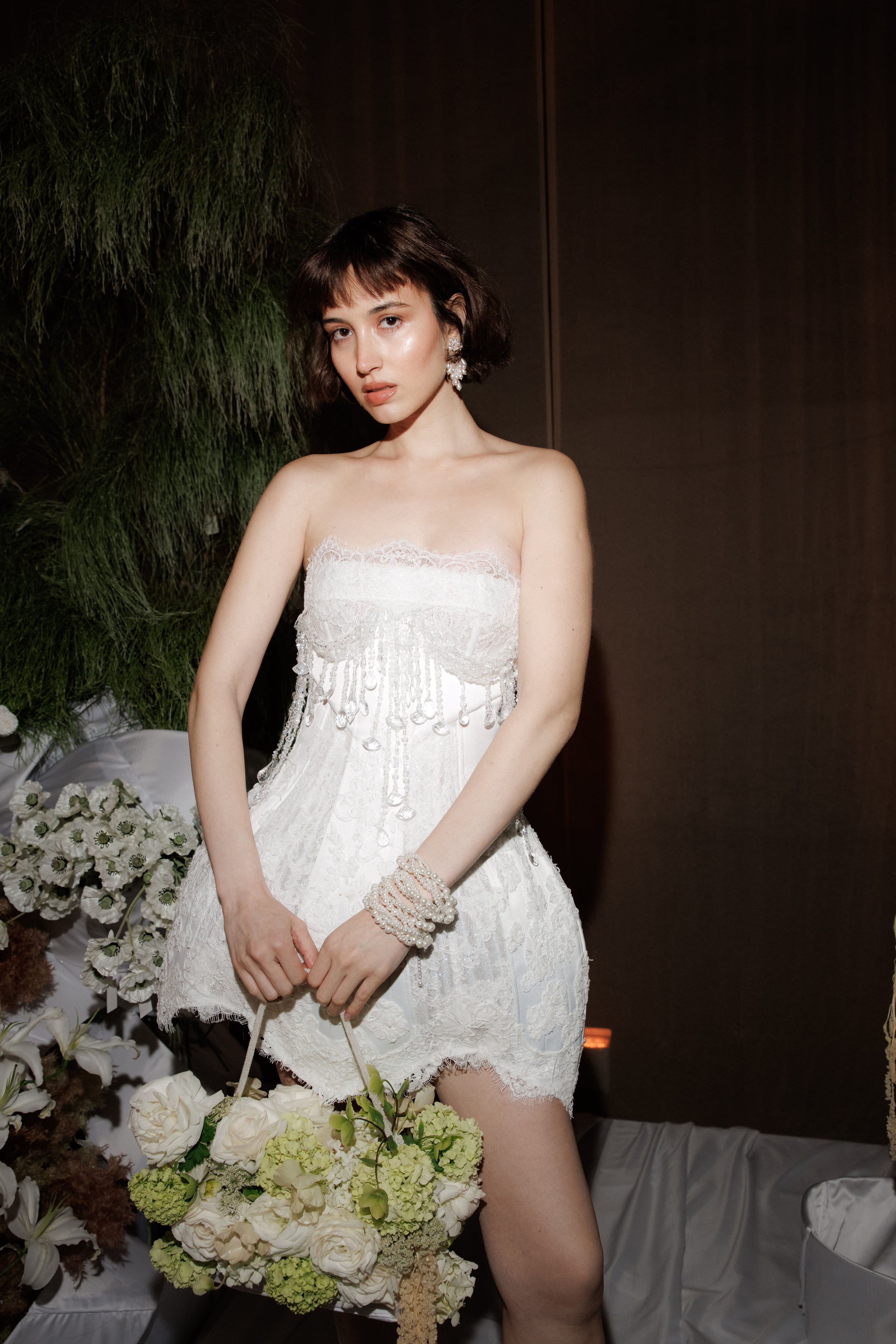 A woman in a strapless white lace mini dress with a beaded crystal fringe at the bodice stands beside a flower arrangement, holding a round bouquet of white garden roses, green hydrangeas and white blooms by a looped handle. Pearl bracelets stack on one wrist, and dark foliage and white poppies frame the scene. This is a wedding flower display in Rethimno, Crete, Greece, shot in soft, low light against a dark backdrop.