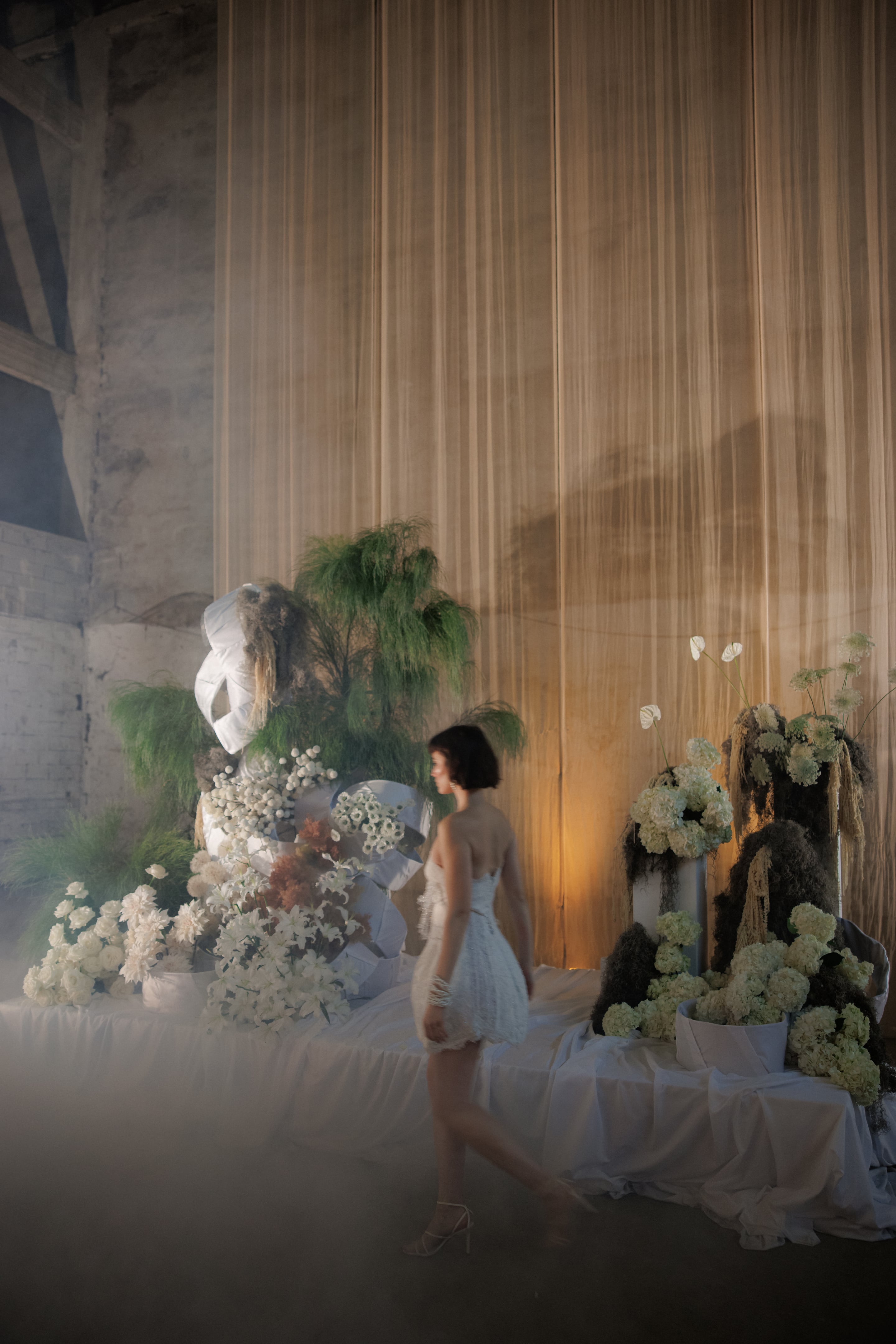 Two large white floral installations rise on a draped white platform, one left and one right, with a woman in a short white strapless dress walking between them past a sheer golden curtain. The left arrangement is built from white lilies, ranunculus, and sprays of tiny white blooms around a tall fern-like green plant, while the right one is heavy with white hydrangeas and white anthurium blooms. Soft haze and warm light create a dreamy, theatrical mood for this wedding flower installation at Rethimno in Crete, Greece.