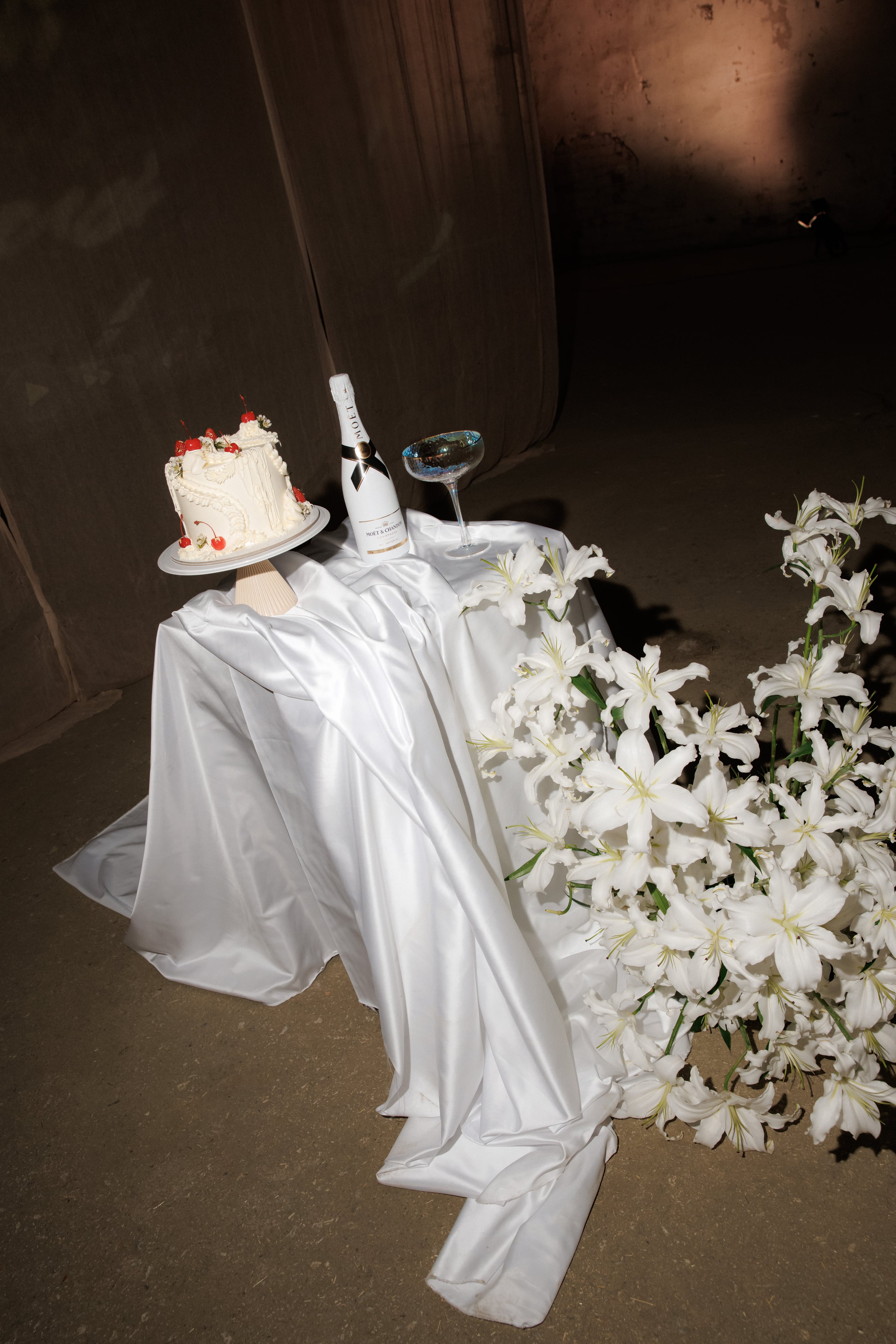 A dramatic display of white lilies, with tall stems of open blooms cascading down beside a draped white satin cloth at a wedding in Rethimno, Crete, Greece. On the cloth sit a small white-iced tiered cake topped with red cherries and tiny white flowers, a bottle of Moët & Chandon ice champagne, and a shallow coupe glass with an iridescent blue sheen. The setting is dim and moody, with dark walls behind, so the white flowers and fabric glow softly.