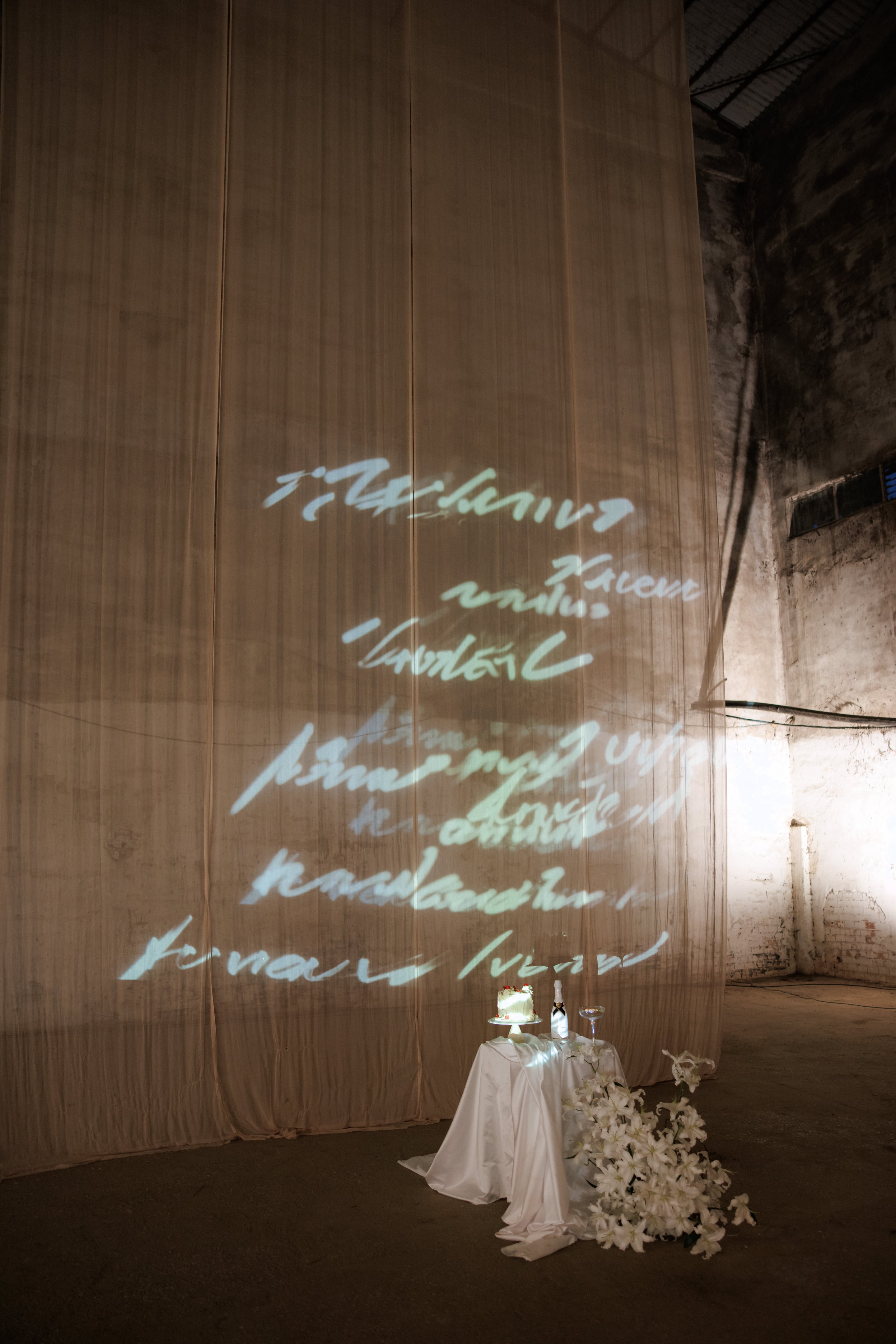 A draped sheer curtain in a bare, weathered industrial hall at Rethimno, Crete, Greece, carries projected handwritten script in soft white light across its full height. At the bottom right, a white-draped table holds a small cake, a champagne bottle and a single flute, with a cascade of white lilies spilling across the concrete floor beside it. The mood is quiet, intimate and airy, set against the crumbling plaster walls.