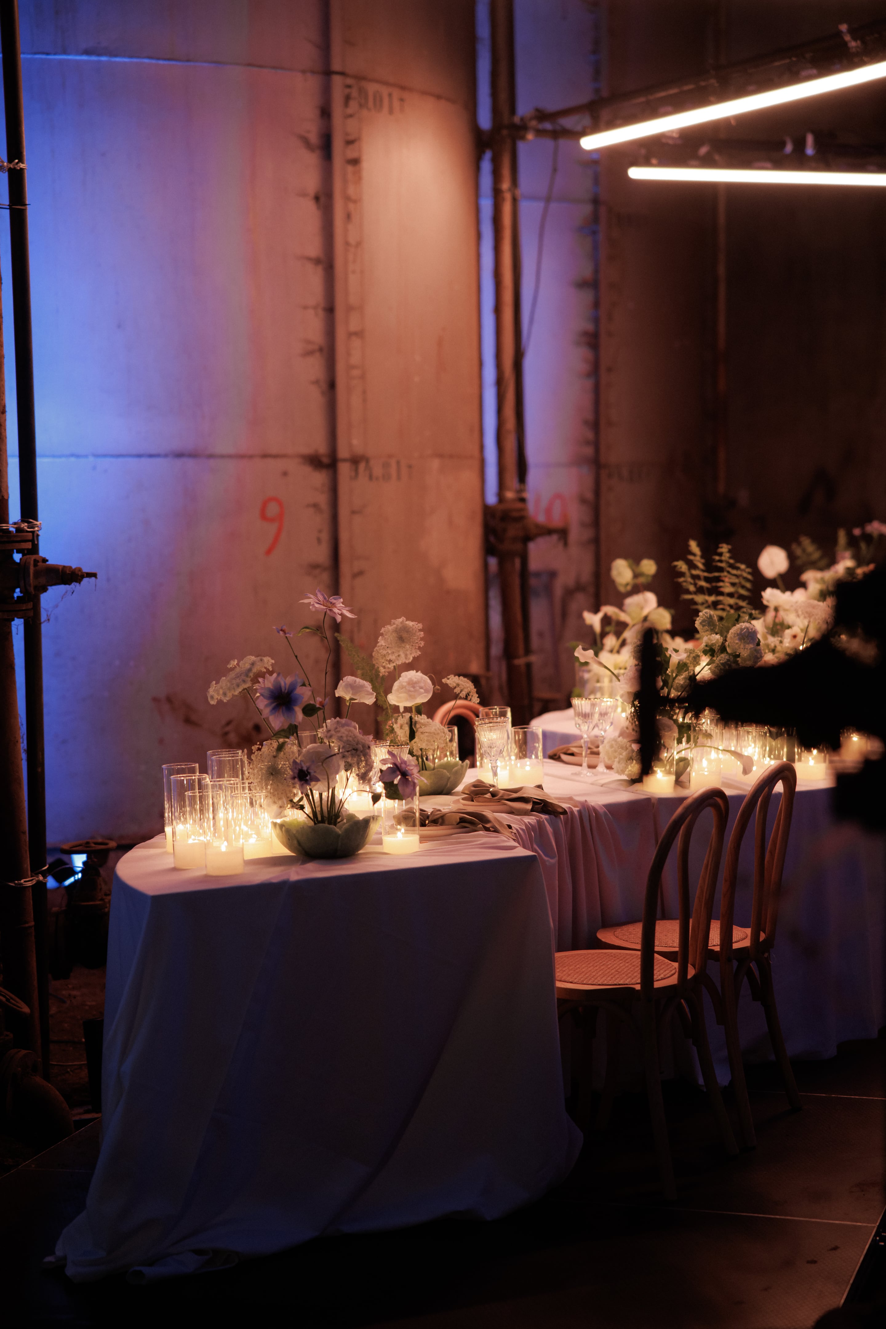 Round tables draped in white linen hold low arrangements of white ranunculus, lacy white hydrangea, and lavender-blue cosmos set in green bowls, with pale fern fronds among them. Clusters of glass votive candles glow in the foreground, and the warm light reflects off the wine glasses. Behind them, weathered concrete walls and rusted pipes are washed in soft blue and pink light, giving this wedding table setting in Rethimno, Crete, Greece a moody, romantic feel.