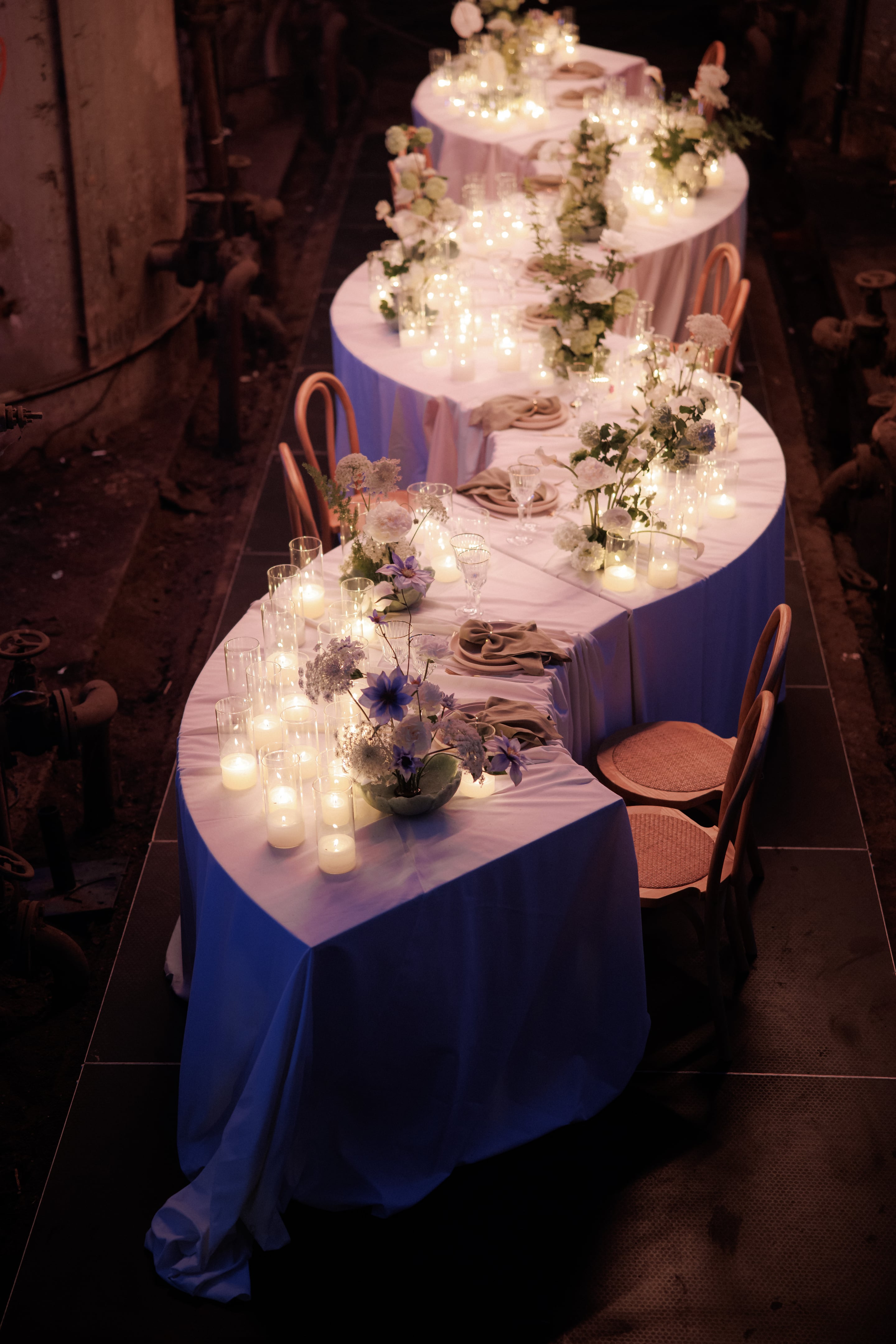 A long, S-shaped wedding banquet table draped in white linen runs diagonally through a dim, industrial space, lined with dozens of flickering pillar candles in glass hurricanes. Low arrangements of white garden roses, hydrangeas, blue clematis and wild carrot flowers sit between folded taupe napkins and bentwood chairs, all warmly lit. This wedding table setting is at Rethimno in Crete, Greece.