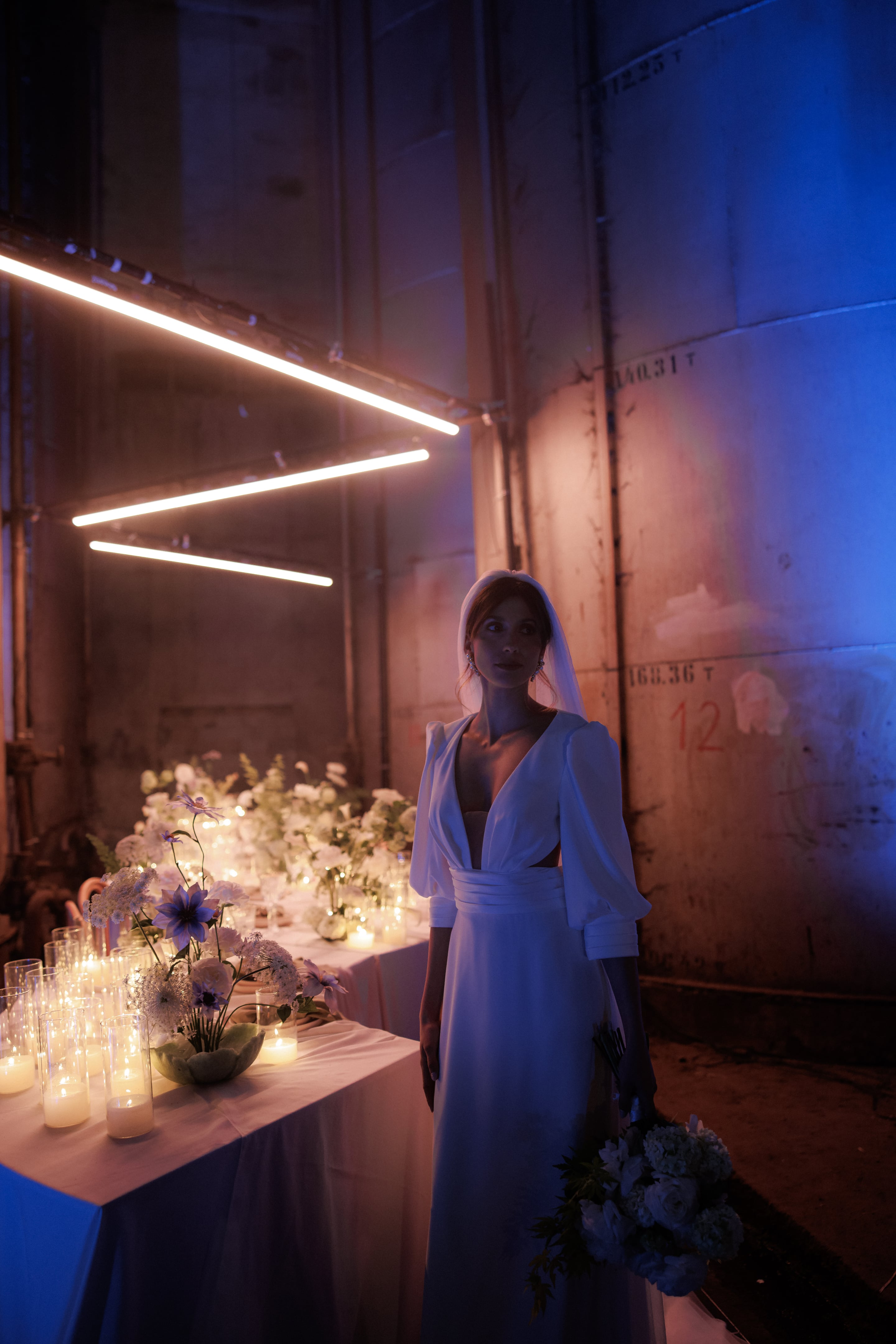 A bride in a white crepe gown with deep V neckline, puffed sleeves and a cinched waist stands in an industrial hall in Rethimno, Crete, Greece, beside a long table of wedding flowers. The table is dressed with white and cream blooms, purple anemones and lacy white umbels, with glass hurricane candles glowing along its edge. Overhead, neon tube lights in pink and warm white meet a blue-lit concrete wall stamped with industrial markings.