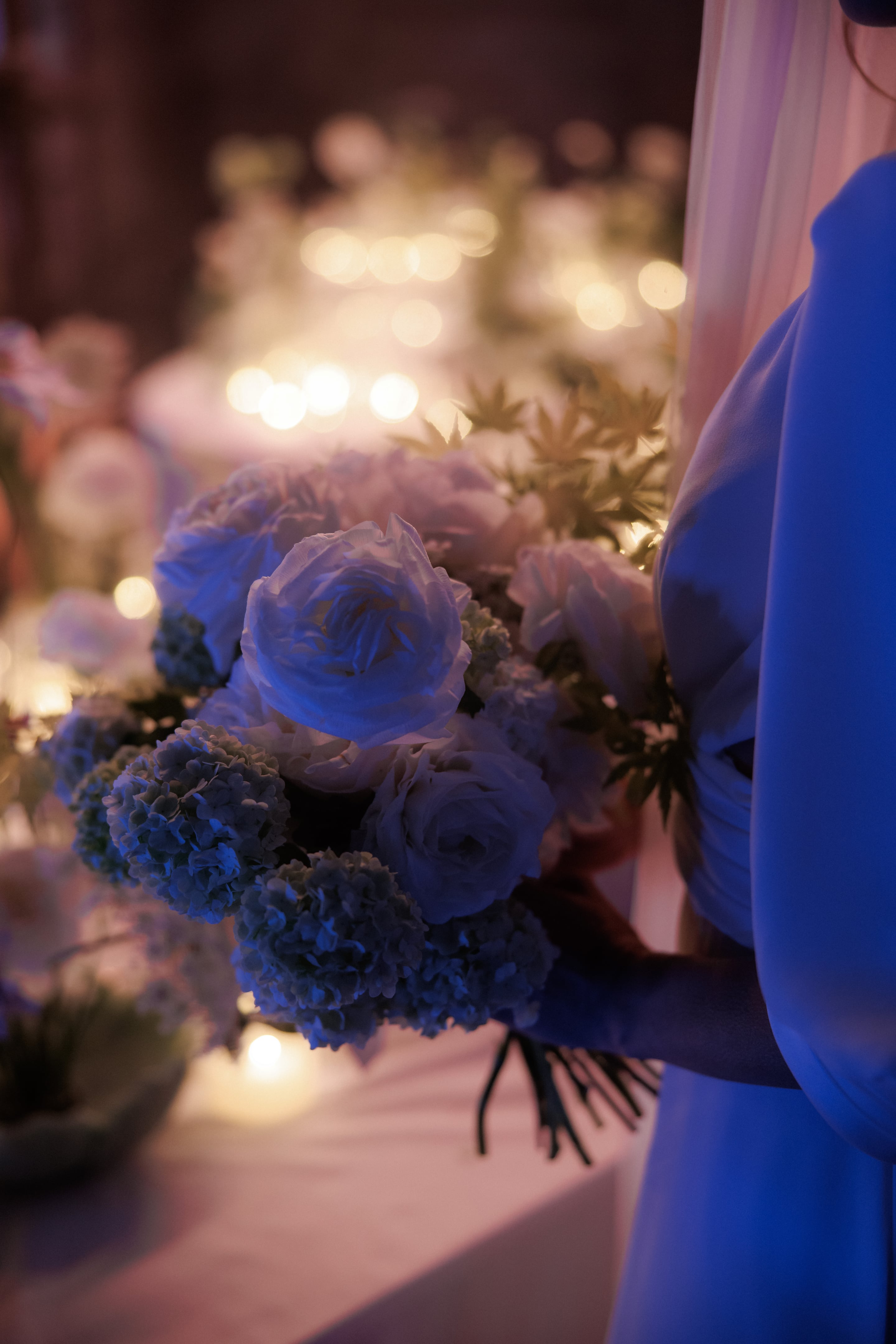 A close view of a wedding floral arrangement in Rethimno, Crete, Greece, lit in cool blue with soft golden bokeh in the background. Pale ranunculus and garden roses sit among clusters of blue hydrangea, with spiky green foliage fanning out behind them. On the right, the edge of a flowing blue gown brushes into the frame, and the warm glowing lights blur gently behind the blossoms, giving the scene a dreamy, evening mood.