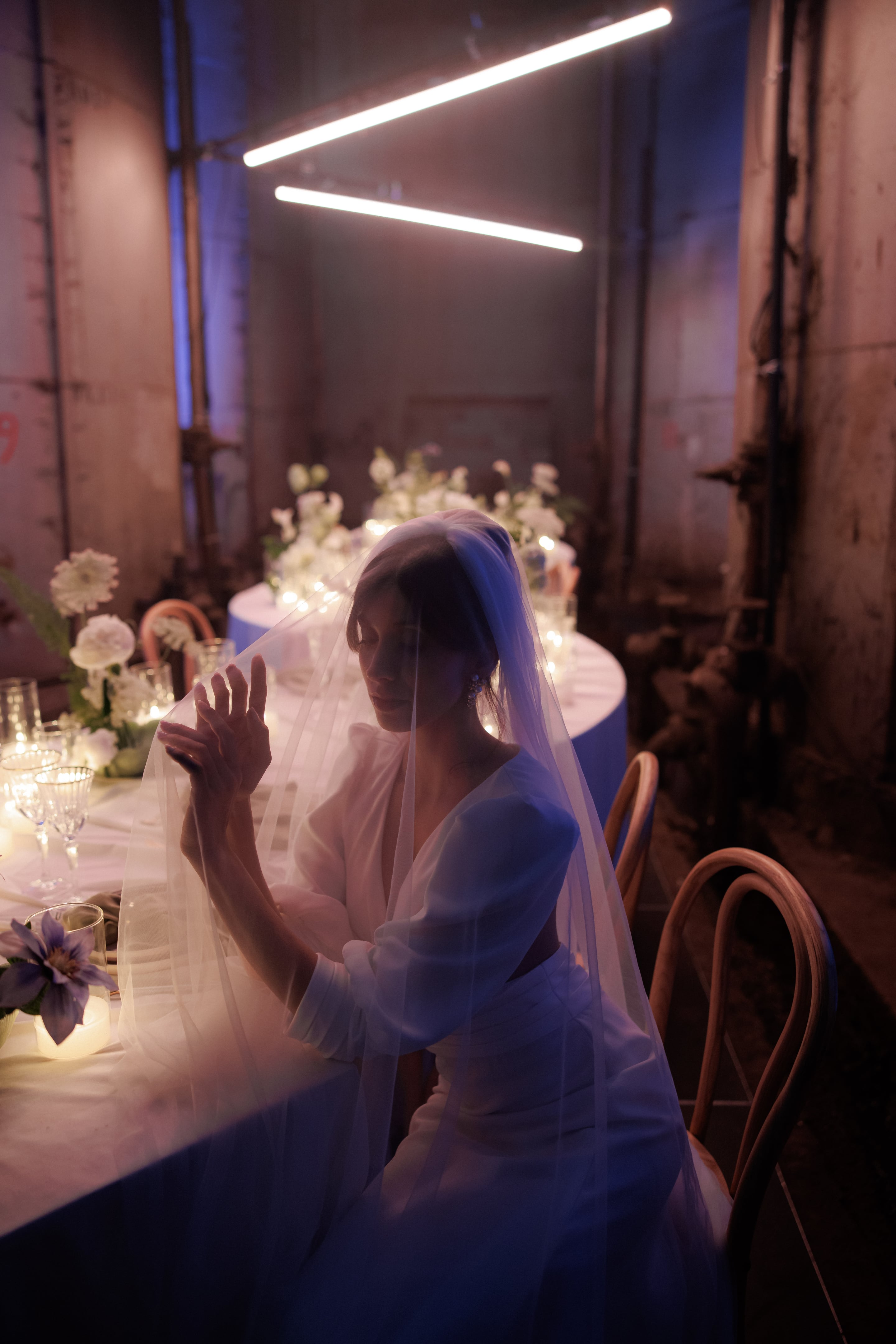 A bride sits in a dim, industrial hall in Rethimno, Crete, Greece, her face softened behind a sheer veil that falls over her shoulders as she lifts one hand. Behind her, a round table dressed in white is lit by strings of tiny candles and set with white garden blooms and clear glassware, with long overhead tube lights glowing above. The mood is quiet, moody and intimate, with blue and pink tones washing over the weathered concrete walls. This is a wedding table styled with white flowers in Greece.