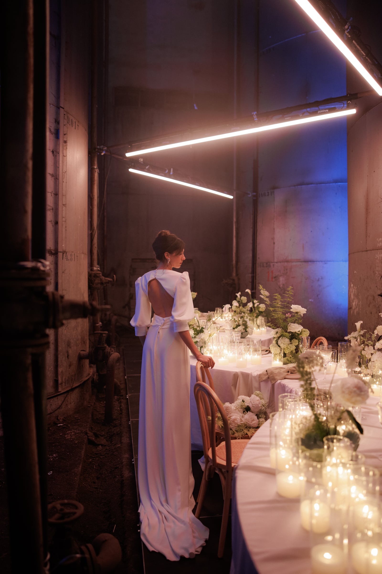 A bride in a long white satin gown with sculpted sleeves and an open back stands beside a long table in a converted industrial hall, her hair in a low bun and her hand resting on a bentwood chair. The table is dressed in white linens and set with clusters of white roses, hydrangeas, calla lilies and fern, with rows of candles glowing in glass holders. This is a wedding table in Rethimno, Crete, Greece, lit by tube lights overhead and a soft blue wash on the tall metal tanks behind her.