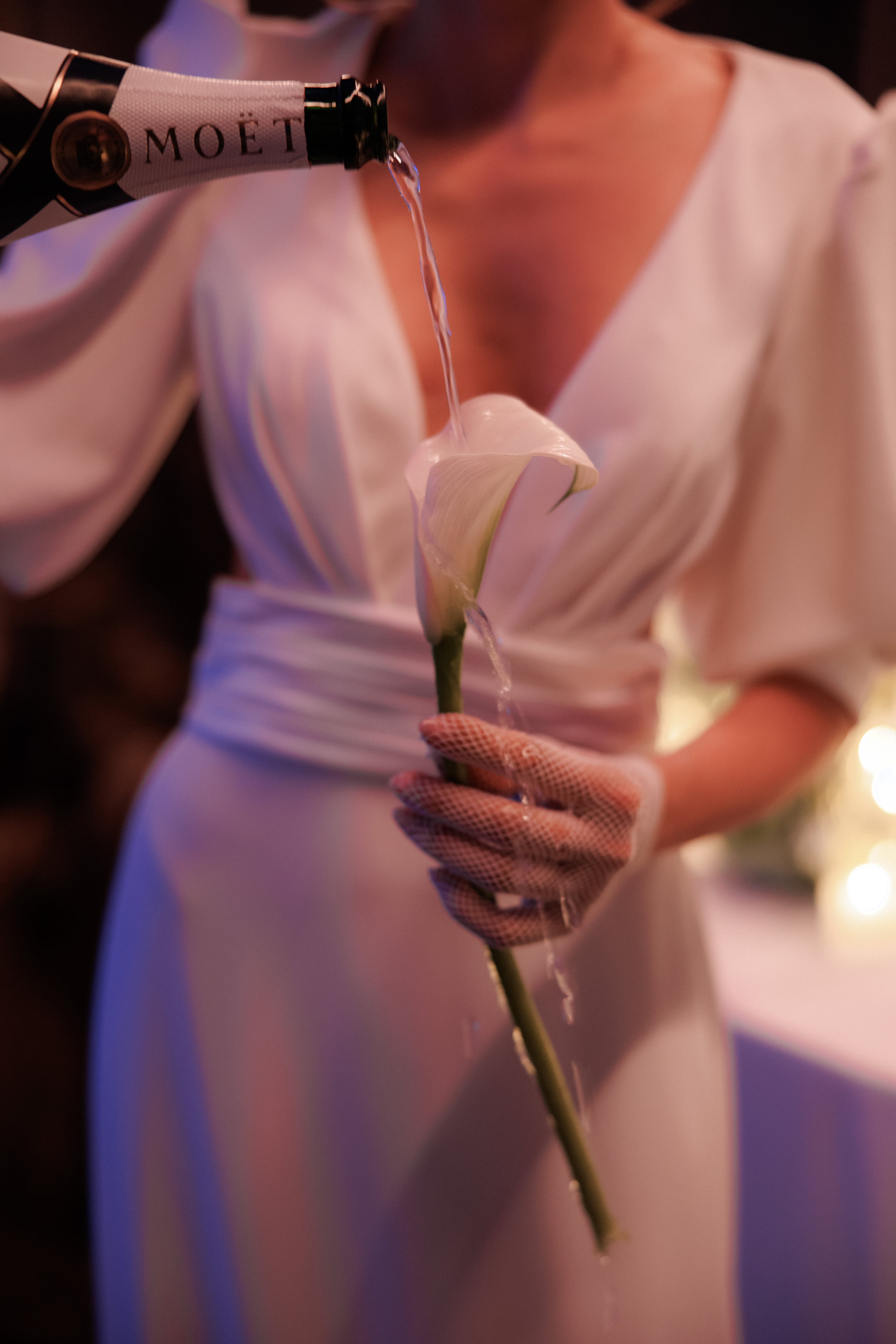 A single white calla lily, its curled cream petals and green stem held in a gloved hand, while Moët champagne is poured over it in a thin stream. The wearer is in a white wrap dress with a deep V-neck and a soft sash, and the warm, low light gives the scene a glowing, intimate mood. This is a wedding flower moment in Rethimno, Crete, Greece.