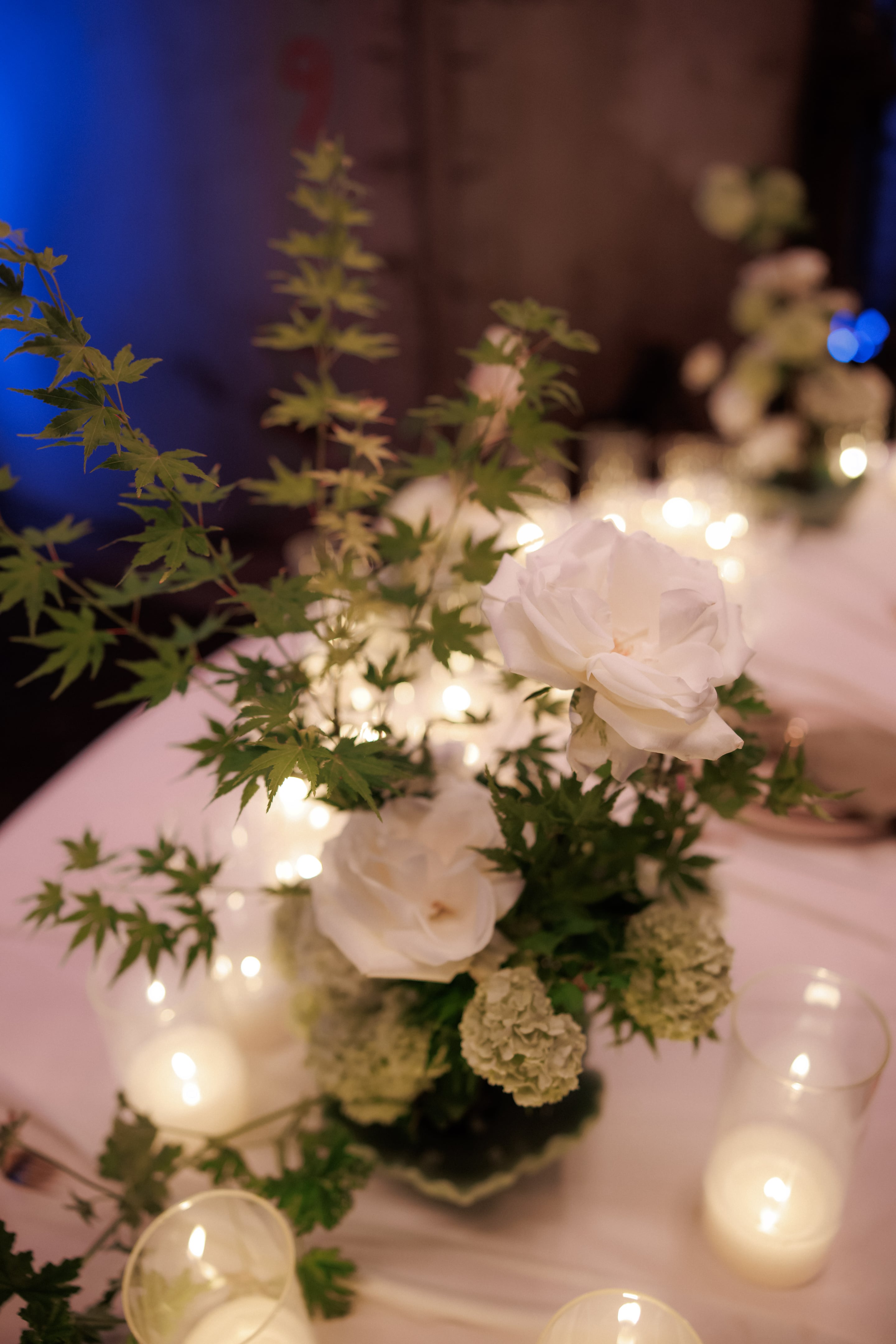 A low table centrepiece of white garden roses and pale green hydrangea heads, threaded through with sprays of fresh green Japanese maple leaves, all glowing in the warm light of candles and fairy lights at a wedding reception at Rethimno Crete in Greece. The white petals and soft bokeh of tea lights in clear glass holders create a calm, romantic mood.