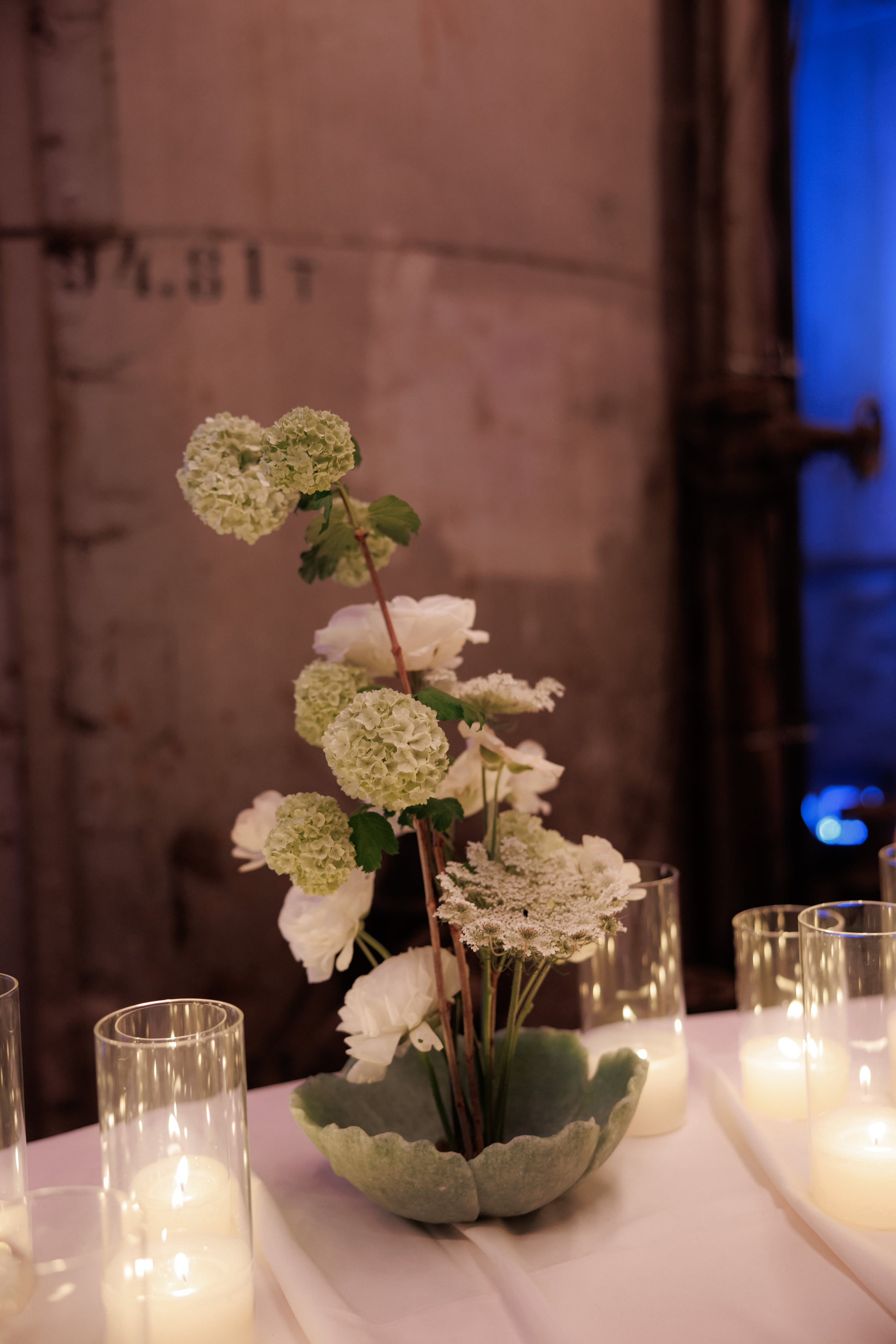 A tall, airy table centrepiece of white and green wedding flowers rises from a scalloped sage-green ceramic bowl, with white ranunculus, green snowball hydrangea, and delicate white Queen Anne's lace spilling over bare stems. Clear glass hurricane candles glow around it on a white tablecloth, set in a dim stone-walled room at Rethimno in Crete, Greece.