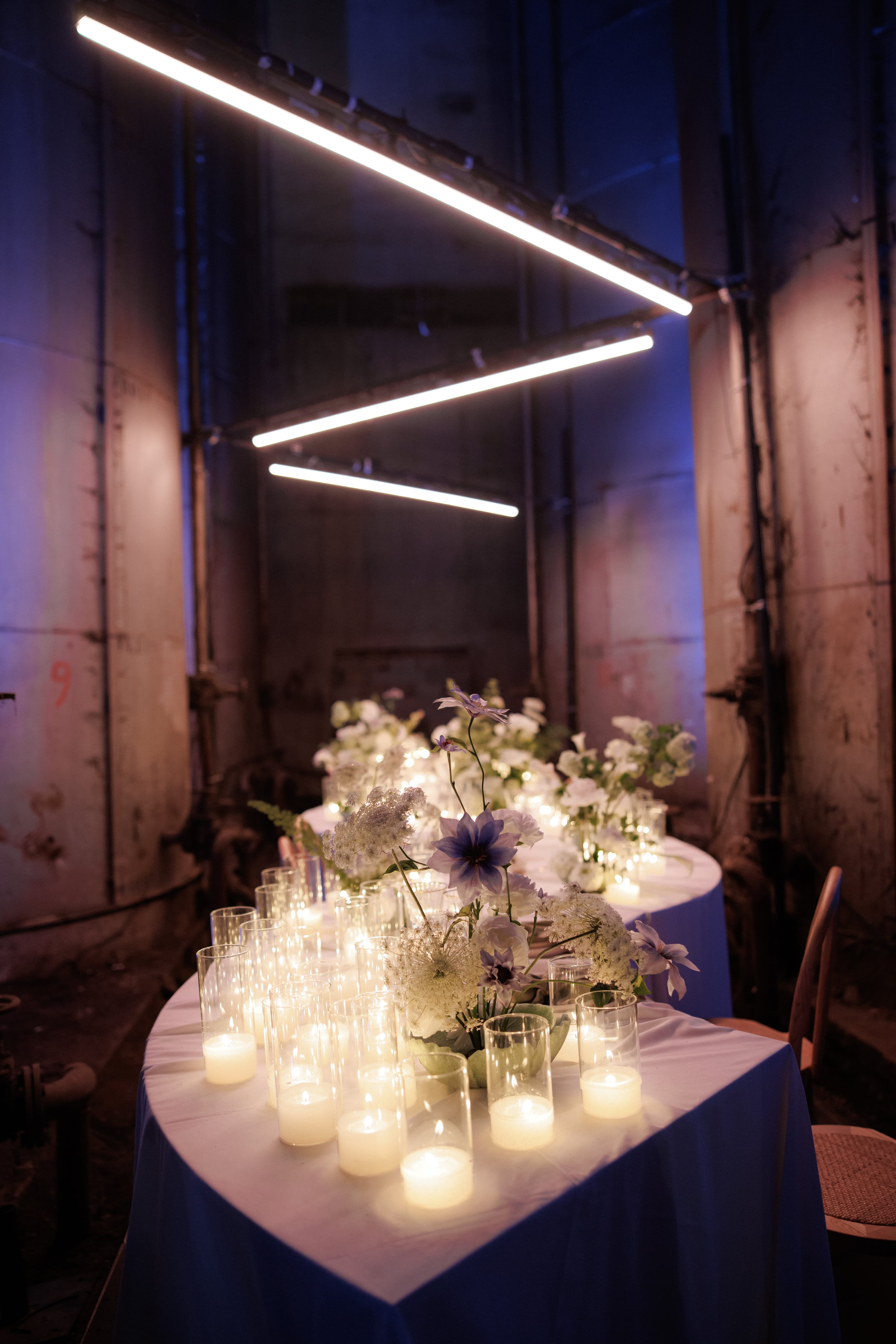A long wedding dinner table in an industrial hall in Rethimno, Crete, Greece, set with low arrangements of white sweet pea and cream blooms, dark violet clematis stems rising above them, and frothy white Queen Anne's lace in the foreground. Clear glasses and tealight candles line the white tablecloth, while three slanted strip lights overhead and the weathered grey metal walls cast a cool blue-violet glow over the scene.