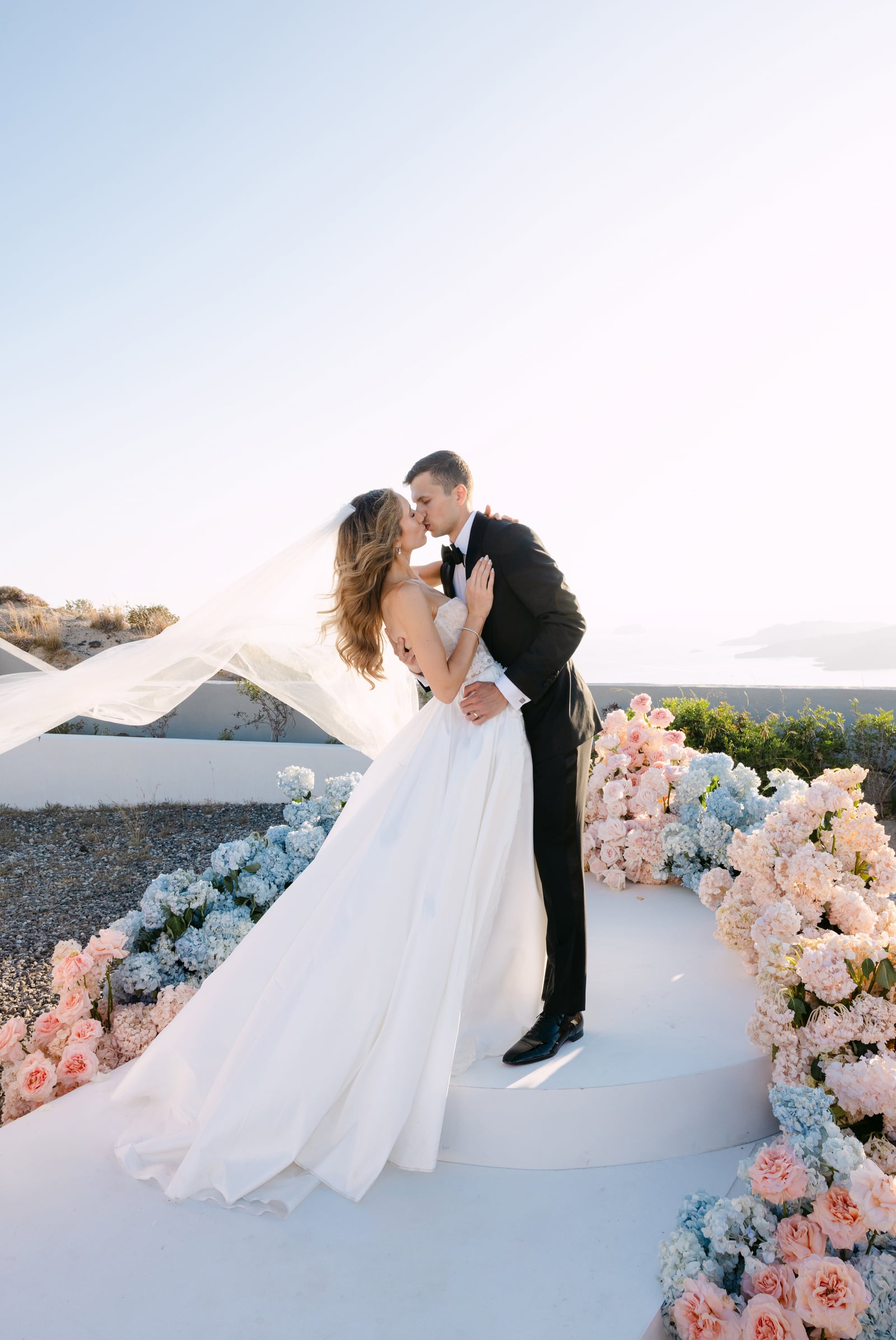 A bride and groom share a kiss on a whitewashed terrace in Santorini, Greece, at Eros Santorini, with a long veil streaming behind her in the breeze. She wears a strapless white satin gown and he a black tuxedo with a bow tie. Along the floor and edges of the terrace, wedding flowers in soft blue hydrangeas, blush garden roses and pale pink peonies spill in lush drifts toward a hazy sea and distant hills under a bright, pale sky.