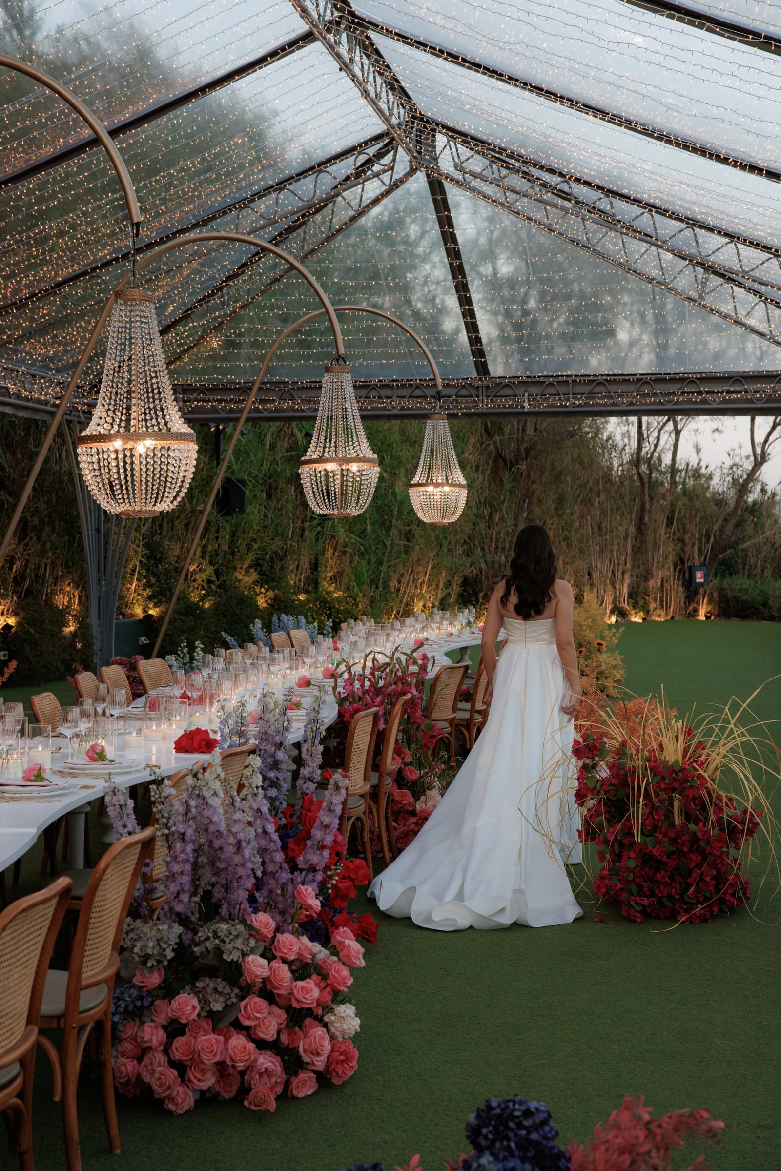 A bride in a strapless white satin gown with a full skirt walks away from the camera, past a long outdoor wedding table set beneath three crystal chandeliers. Along its edge, tall purple delphinium spires rise above drifts of pink garden roses, and clusters of deep red bougainvillea and tall grasses frame her path on the green lawn. This is a wedding table at Island Athens Riviera in Greece, with glassware and candles glowing in the dusk.